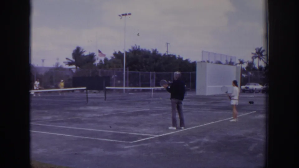 1973 Naples, Florida home-movie frame with adult tennis players on a clay court, including a foreground man holding a racket near court lines.