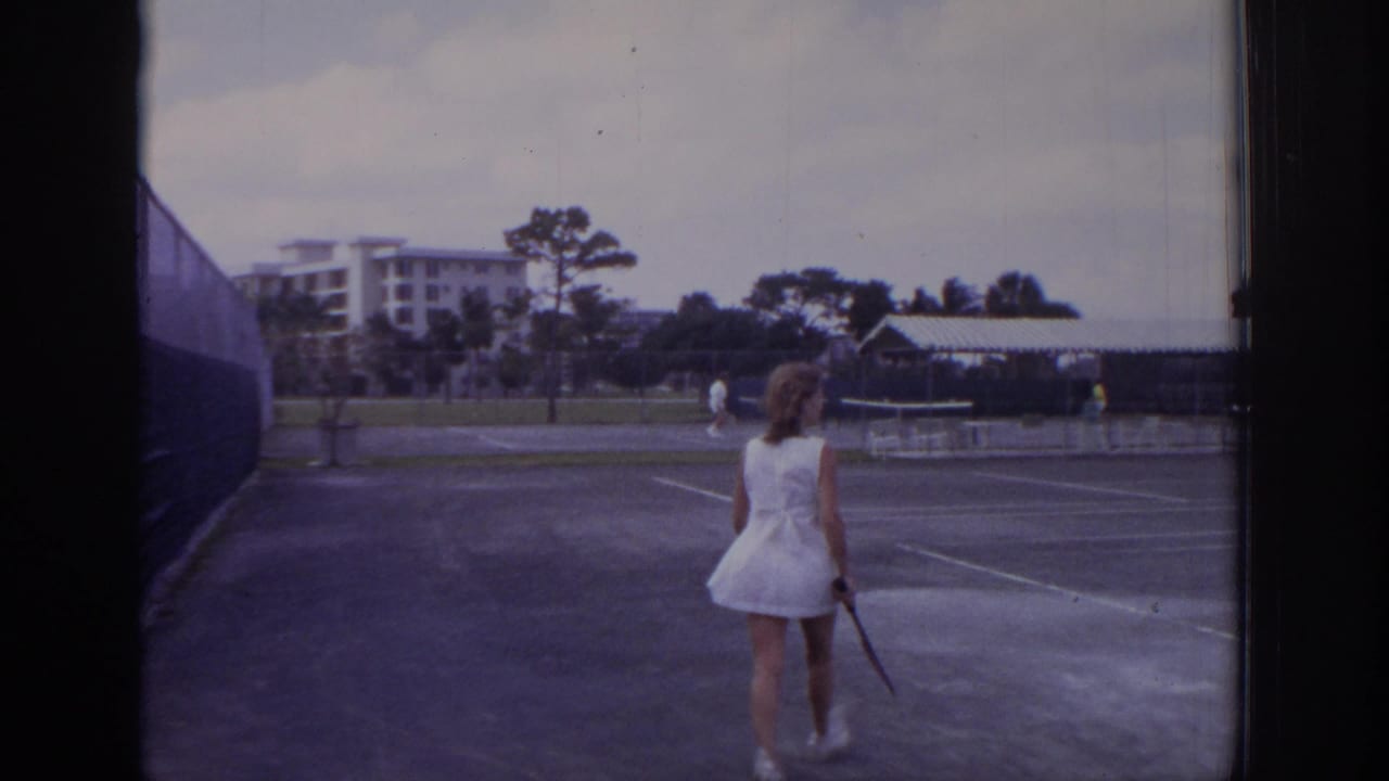 1973 Naples, Florida home-movie frame showing a woman in a white tennis dress walking across a resort court with other players beyond.