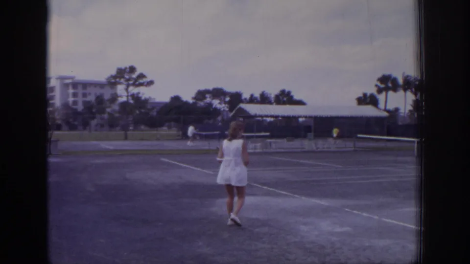 1973 Naples, Florida resort tennis scene with a woman walking across the court and adjacent play visible under a cloudy sky.