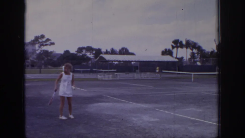 1973 Naples, Florida frame of a woman paused on a resort tennis court, holding a racket with palms and low buildings behind.