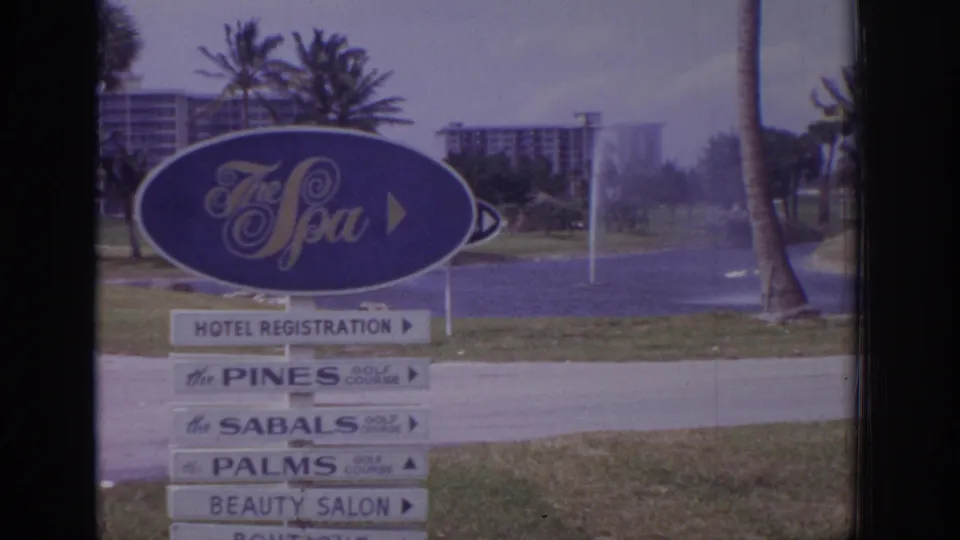 1973 Naples, Florida 8mm frame showing The Spa entrance sign, directional boards, palms, lawn, fountain spray, and distant buildings.