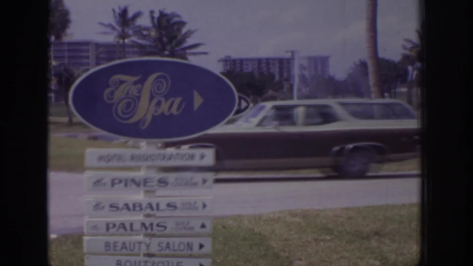1973 Naples, Florida 8mm frame with a station wagon moving past The Spa sign, stacked wayfinding signs, palms, and resort buildings.