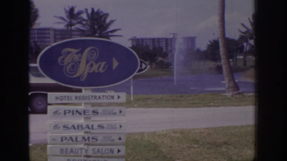 1973 Naples, Florida 8mm frame showing The Spa resort sign, stacked lodging and salon signs, palms, fountain spray, and a partial car.