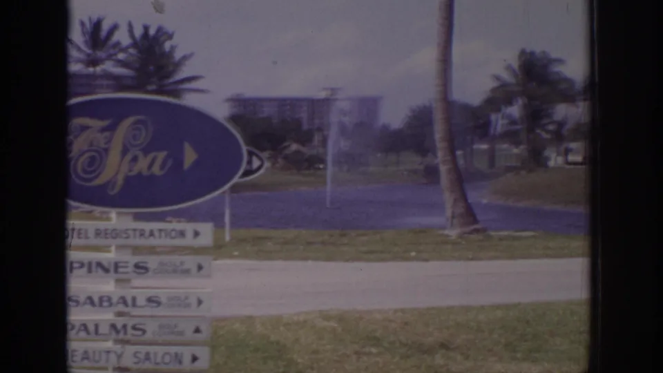 1973 Naples, Florida 8mm frame with the partly cropped The Spa sign, directional boards, grass verge, palms, and distant resort buildings.