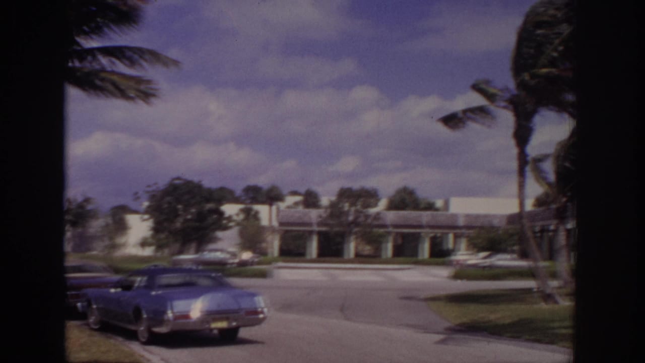 1973 Naples, Florida home-movie frame showing a white building, parked cars, lawns, and palm trees bending in strong wind.
