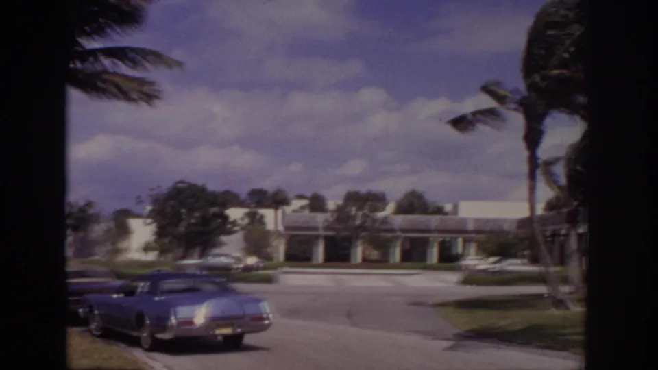 1973 Naples, Florida home-movie frame showing a white building, parked cars, lawns, and palm trees bending in strong wind.