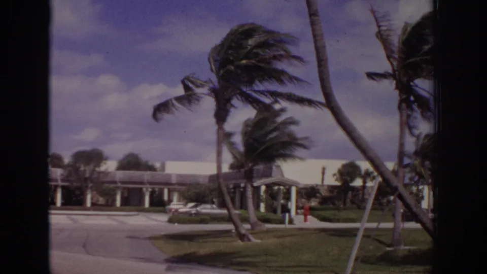 1973 Naples, Florida 8mm frame showing a driveway, white building, covered entrance, and palms leaning in heavy wind.