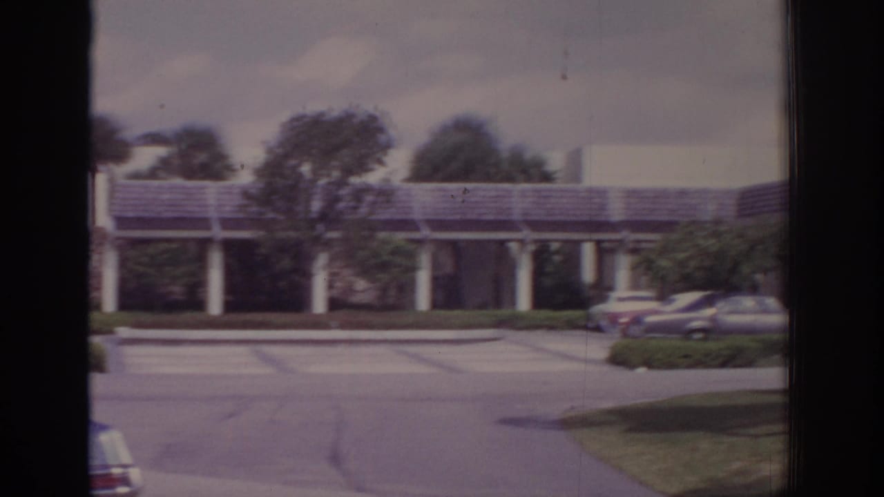 Low building with columns, driveway, parked cars, and wind-blown trees.