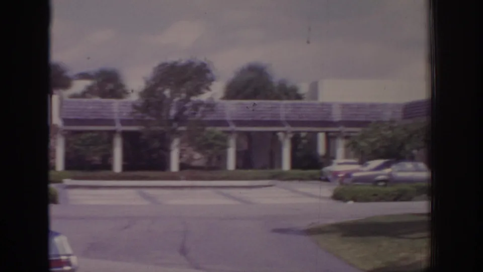 1973 Naples, Florida view of a mid-century style building, driveway, parked cars, and trees moving in heavy wind.