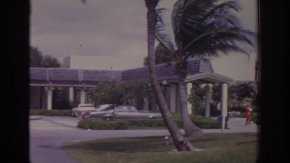 1973 Naples, Florida frame showing leaning palms, a covered building entrance, parked cars, lawn, and a person in red near the walkway.