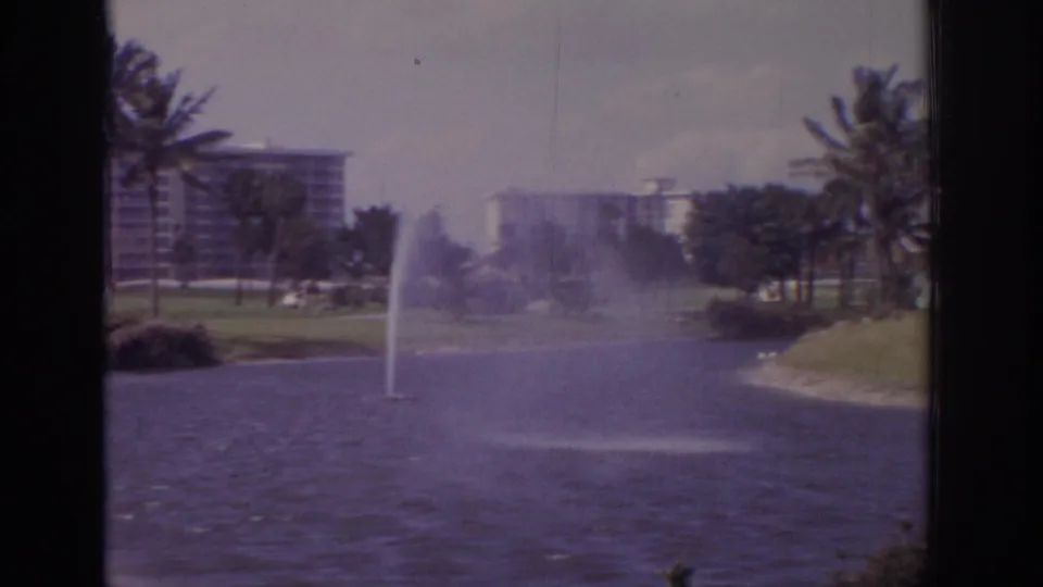 1973 Naples, Florida frame showing a fountain bent by wind over dark lake water, with palms, lawn, and a distant apartment block.