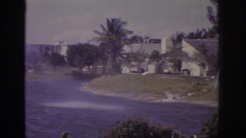 1973 Naples, Florida 8mm frame of a lake fountain in wind, with palms, landscaped grass, shrubs, and white low-rise buildings beyond.