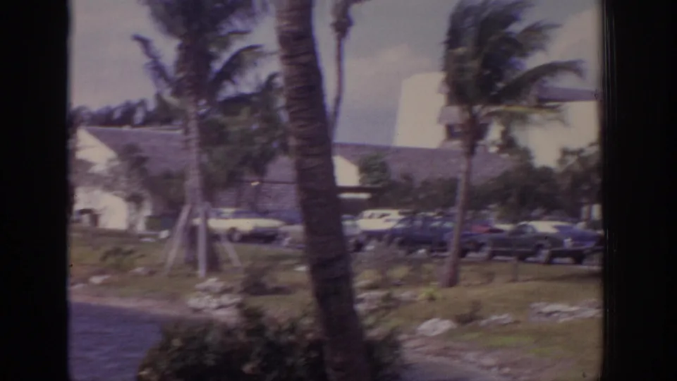 Windy 1973 Naples resort scene with palms, lawn, low hotel buildings, and early-1970s cars in the parking area.