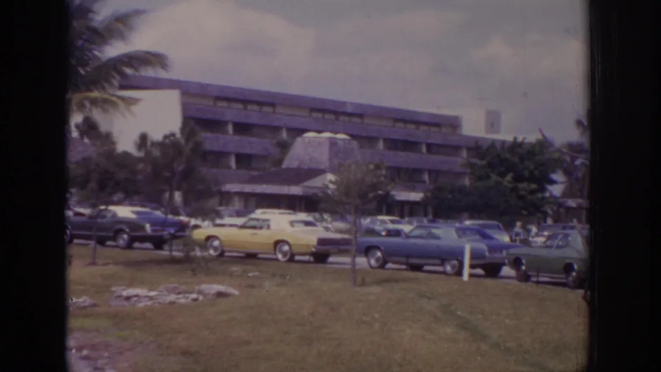Wide 1973 Naples resort view showing classic cars on grass, a yellow sedan, a blue coupe, palms, and a hotel building.