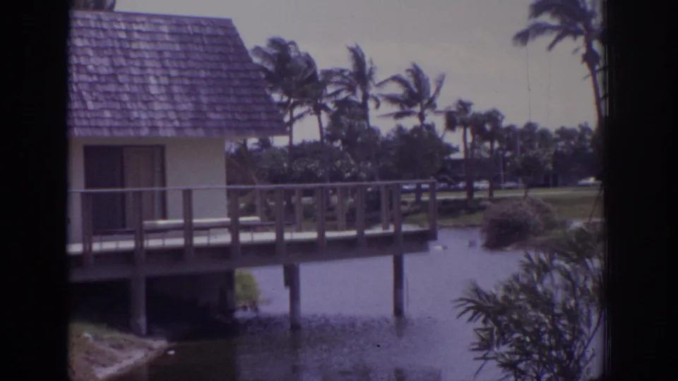 1973 Naples, Florida home-movie frame showing a small stilted structure over calm water with wooden deck railings and palm landscaping in the background.