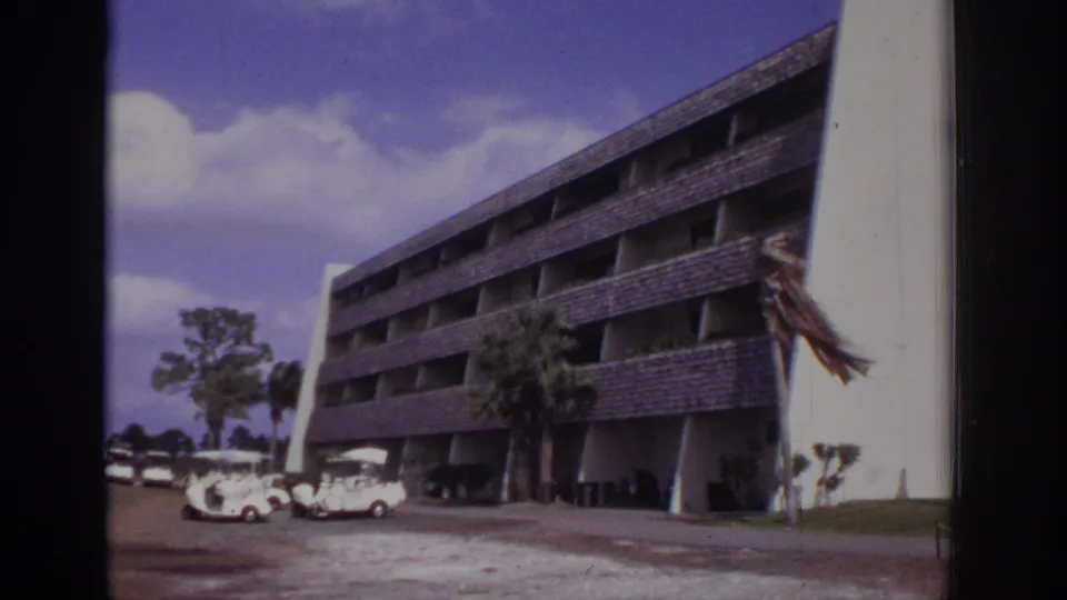 1973 Naples, Florida home-movie frame showing a large resort-like multi-story building with a sloped roof, palm trees, and small parked carts near the entrance area.