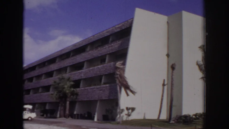 1973 Naples, Florida frame showing the side of a large modern resort-style building, with a dark sloped roof, white end wall, palm trees, and carts parked below.