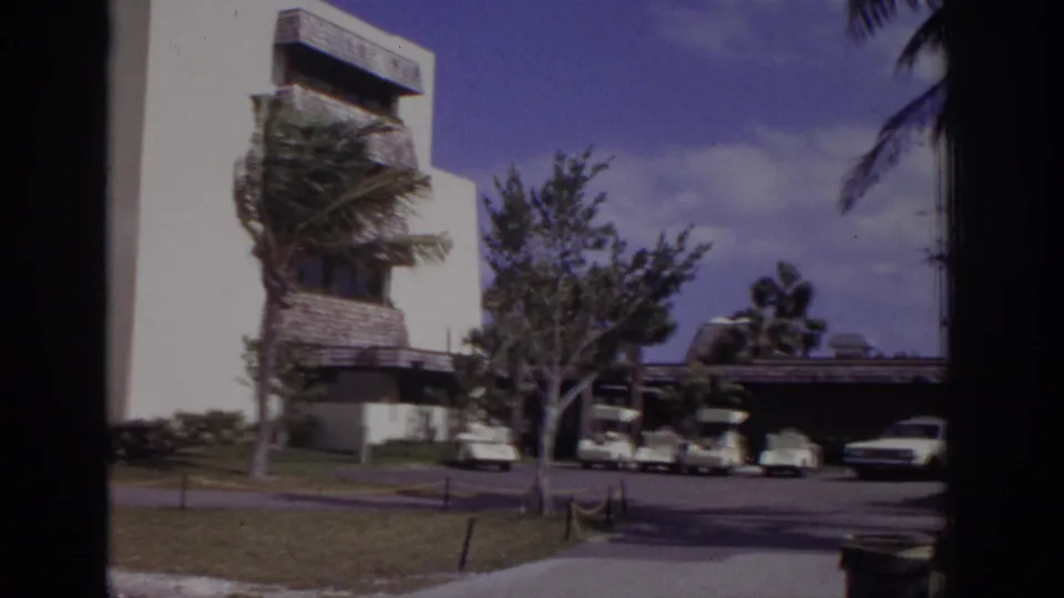 1973 Naples, Florida 8mm view of a building edge, palm, parking area, golf carts, cars, and tropical landscaping.