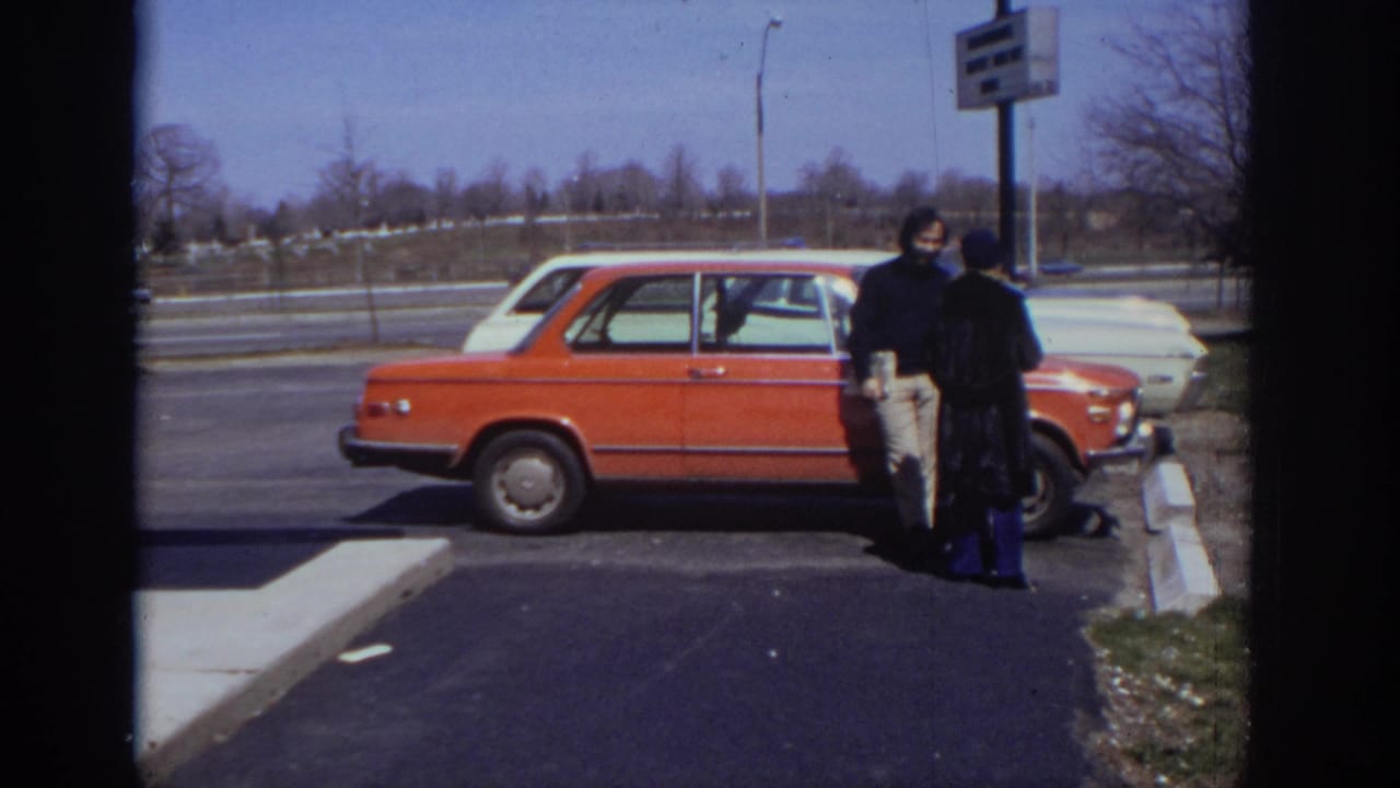 Two adults stand near an orange car in a parking lot beside a road.