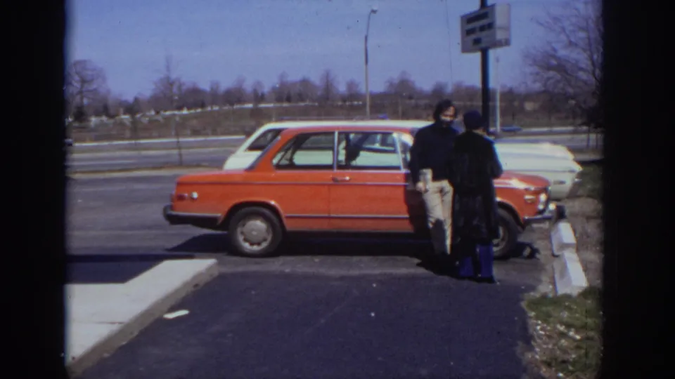 1973 Philadelphia 8mm frame showing two adults near an orange sedan in a sunlit parking lot with road traffic beyond.