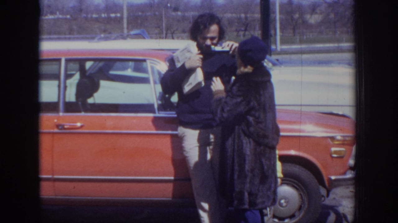 1973 Philadelphia 8mm frame of a man with a newspaper talking to a woman in a dark coat beside an orange sedan.