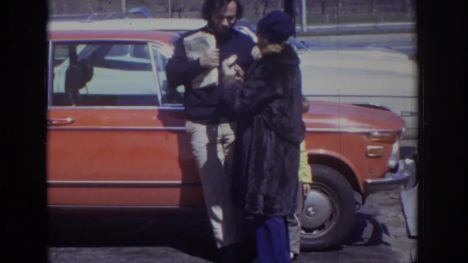 1973 Philadelphia 8mm frame showing the couple beside the orange sedan, with the newspaper visible and road traffic in back.