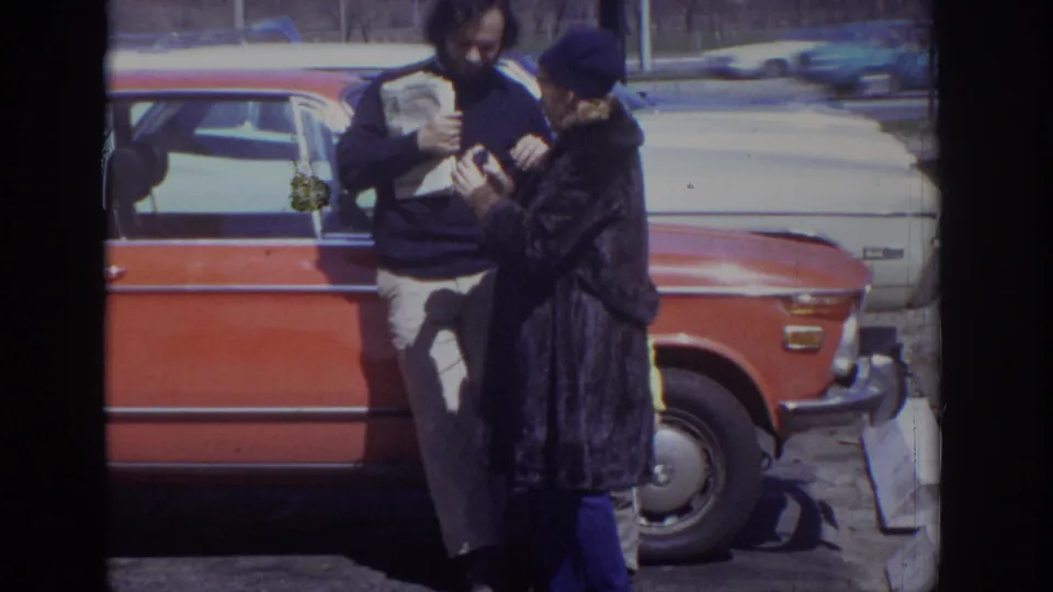1973 Philadelphia 8mm frame of a woman in a dark coat gesturing toward a man with a newspaper beside an orange sedan.