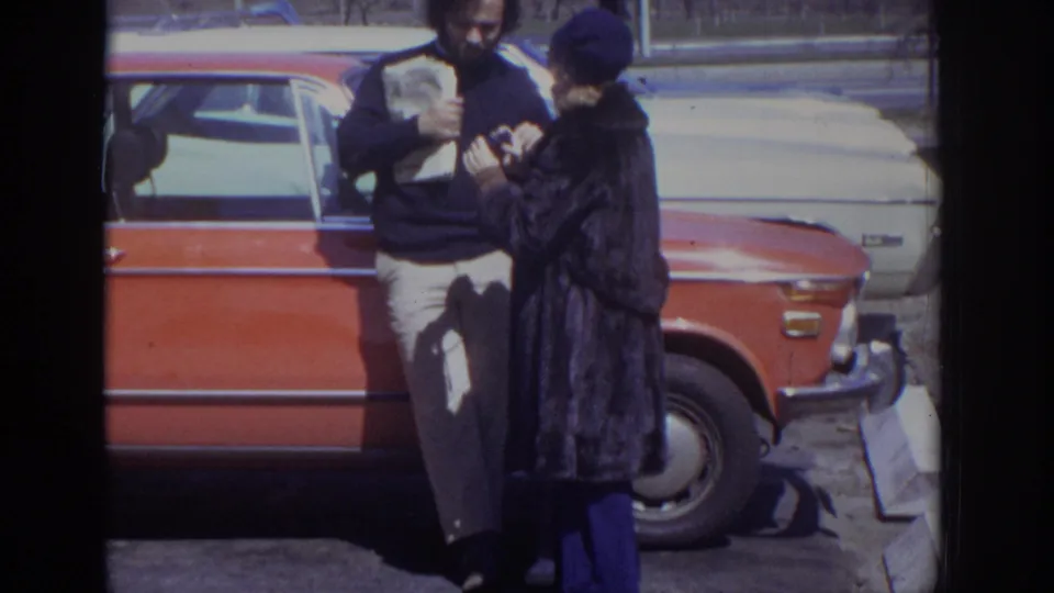 In 1973 Philadelphia home-movie footage, a man and woman stand by a red station wagon and look at something in their hands.