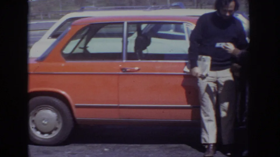 A man in a dark top and light trousers stands beside the red station wagon while the woman is mostly obscured at the edge.
