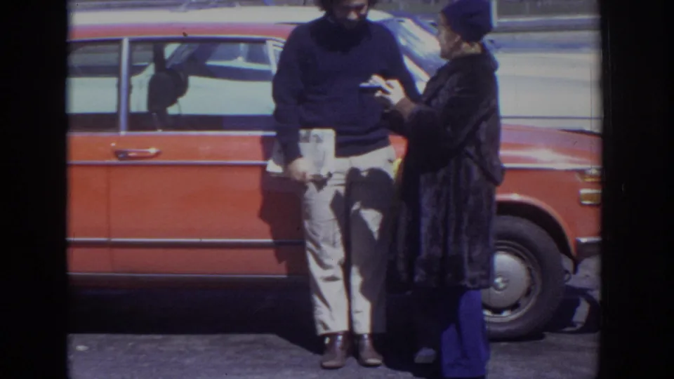 Beside the red station wagon, the woman appears to show a small object to the man during their brief exchange.