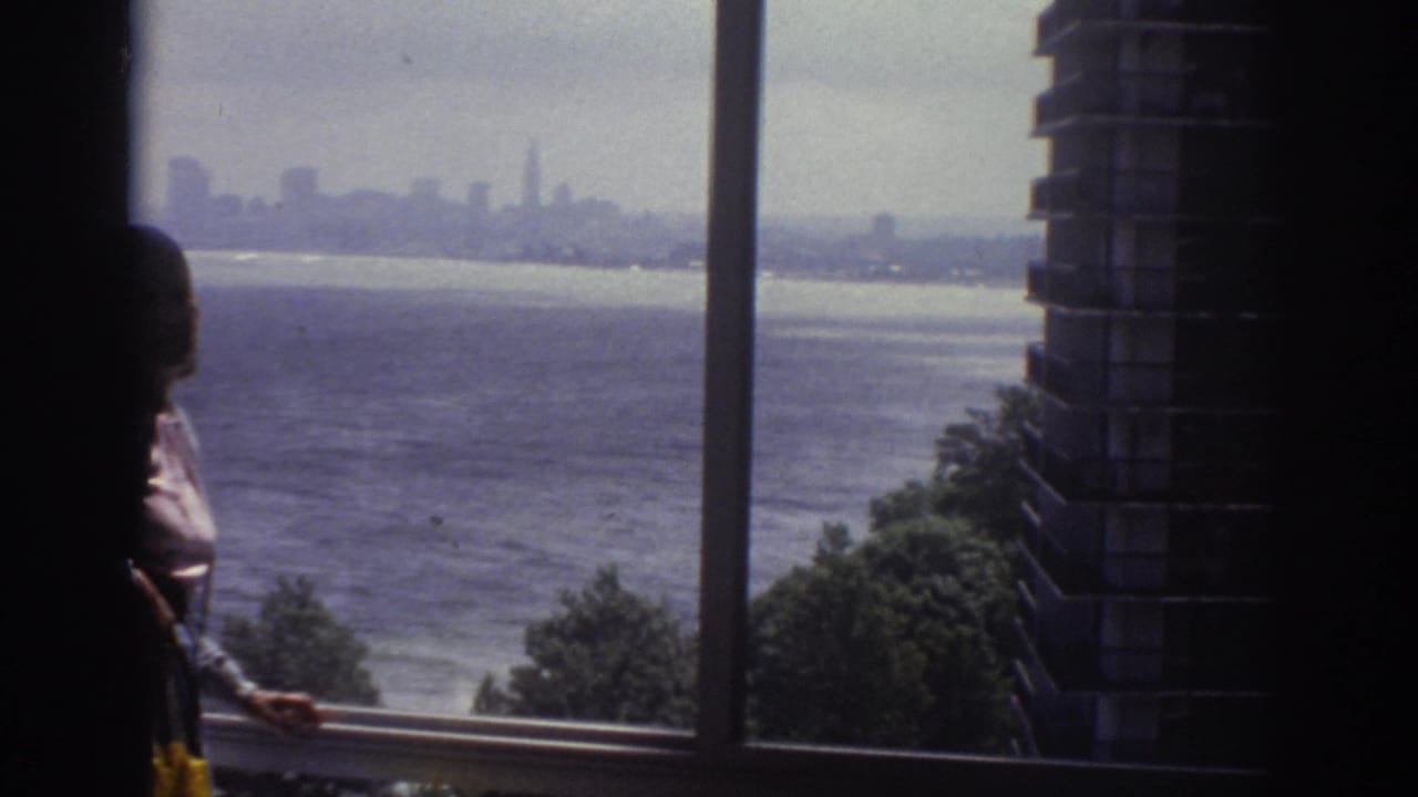 1974 Cleveland home-movie frame with a silhouetted person at a window looking toward water, shoreline trees, and a distant skyline.