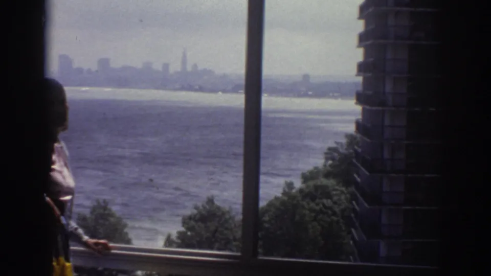 1974 Cleveland home-movie frame with a silhouetted person at a window looking toward water, shoreline trees, and a distant skyline.