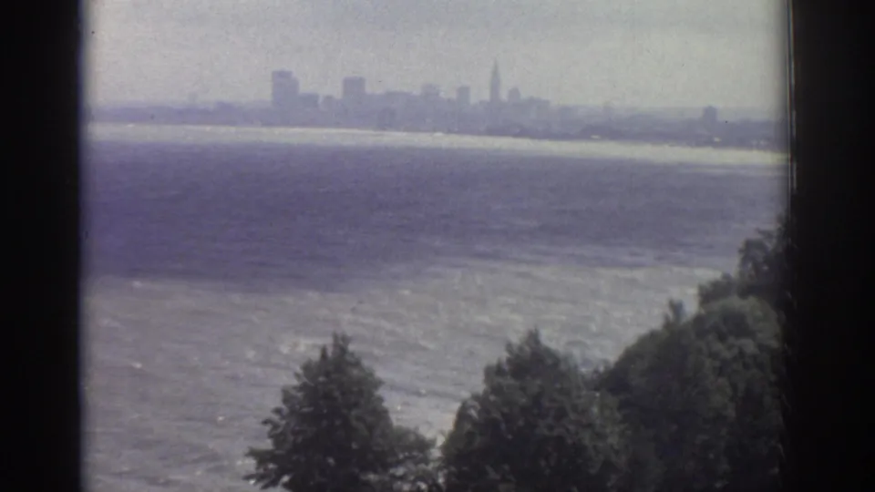 1974 Cleveland view across a broad water surface toward a distant skyline, framed by a dark window edge and shoreline trees.