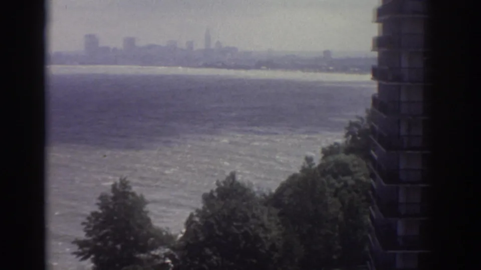 1974 Cleveland home-movie view with water in the foreground, shoreline trees, a nearby high-rise facade, and a distant skyline.