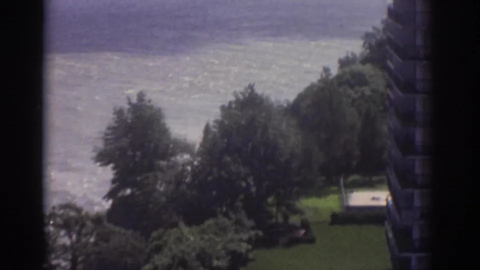 1974 Cleveland frame looking down toward shoreline trees, a green lawn, and bright water beyond a nearby building edge.