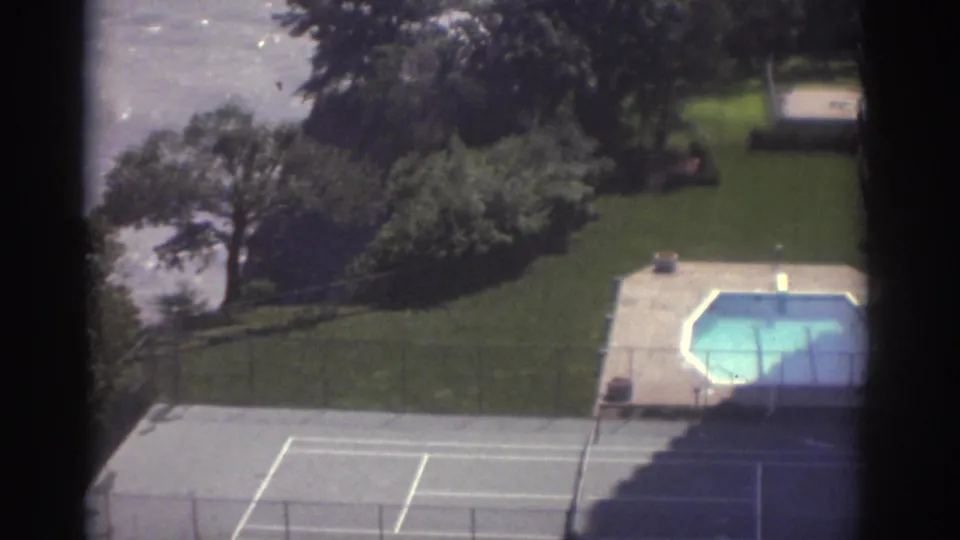 1974 Cleveland 8mm view over a lakeside property with a tennis court, octagonal pool, lawn, trees, and lake water beyond.