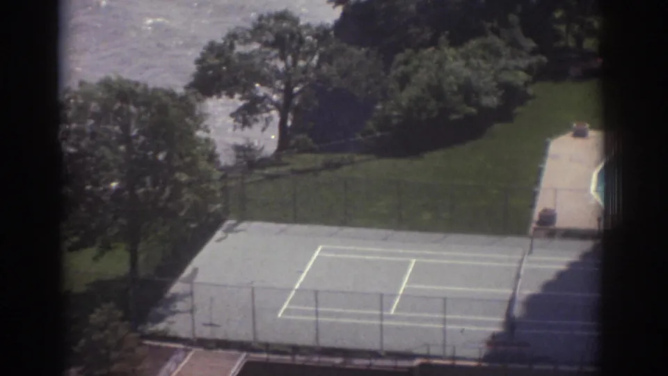 1974 Cleveland 8mm frame showing the tennis court below, bordered by lawn, trees, fencing, and lake water in the background.