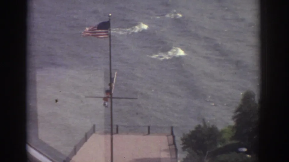 1974 Cleveland 8mm frame with an American flag on a tall pole near choppy lake water, a paved waterfront edge, and nearby trees.