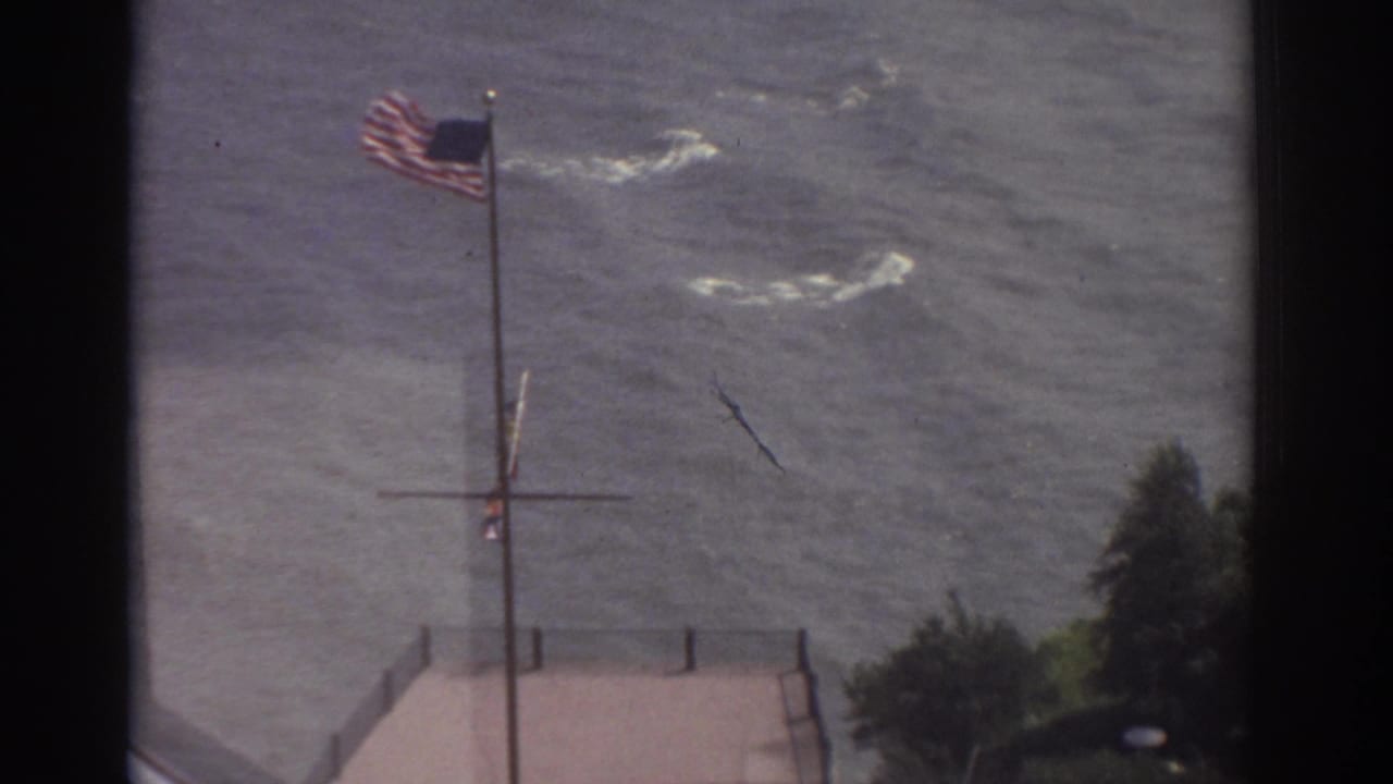 1974 Cleveland waterfront view with an American flag blowing above rippling water and a pier edge in soft 8mm color.
