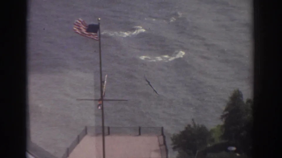 1974 Cleveland waterfront view with an American flag blowing above rippling water and a pier edge in soft 8mm color.