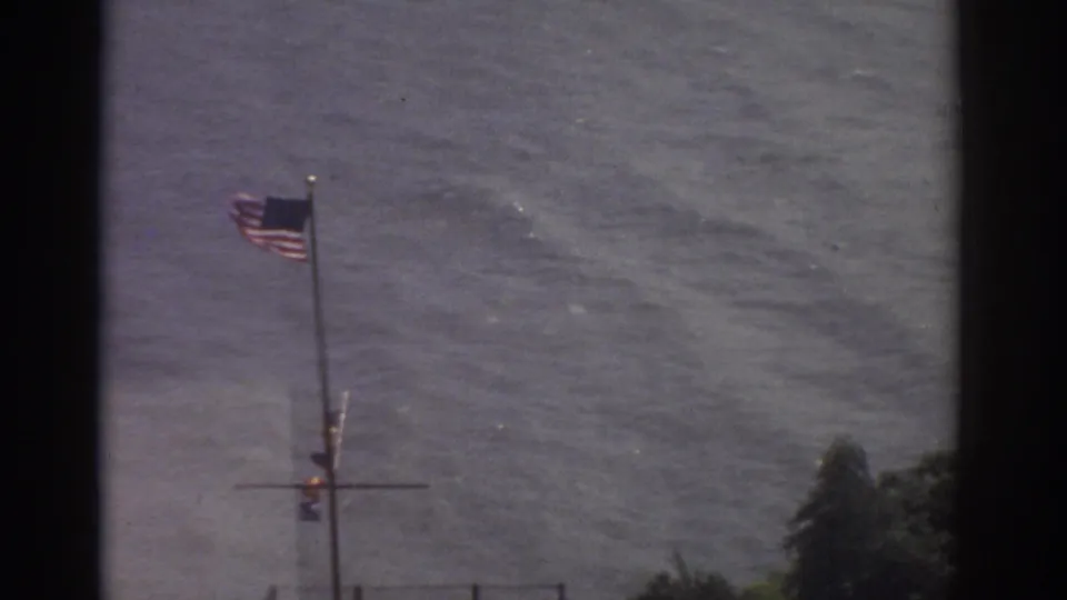 Handheld 8mm frame of an American flag above Cleveland waterfront water, with trees and pier details near the lower edge.