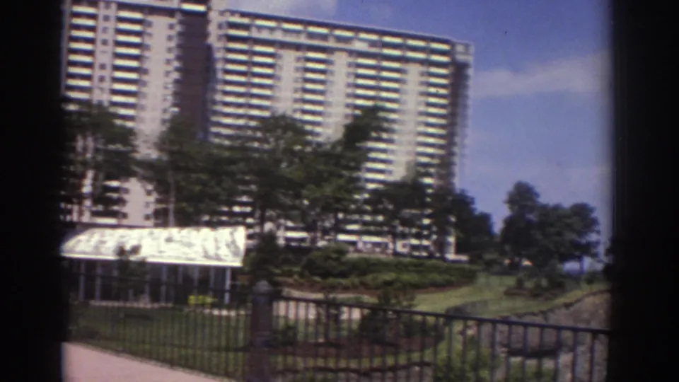 1974 Cleveland 8mm view of a high-rise apartment building near railing, trees, landscaped slope, and open water.