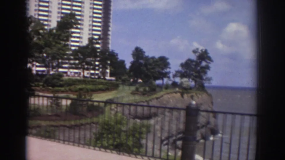 1974 Cleveland frame looking past a metal railing toward landscaped grounds, a high-rise tower, trees, and water.