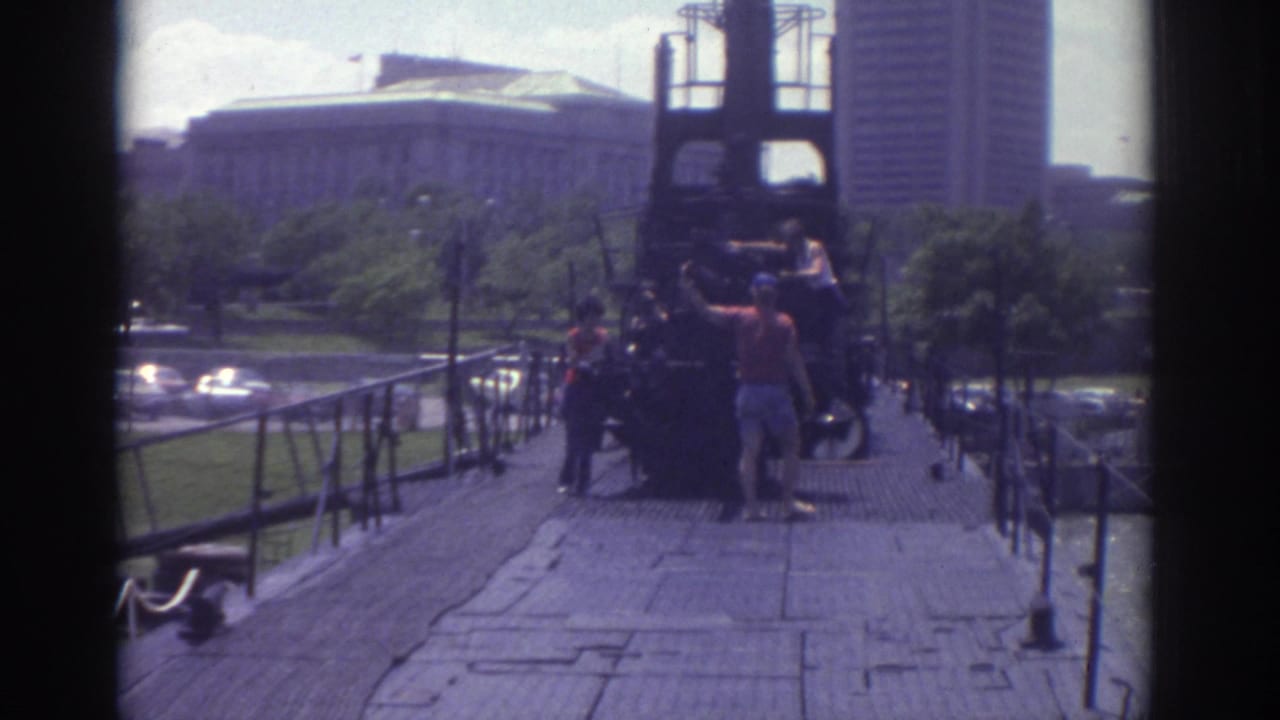 1974 Cleveland home-movie frame showing people on a wooden walkway near a dark structure with downtown buildings in the background.