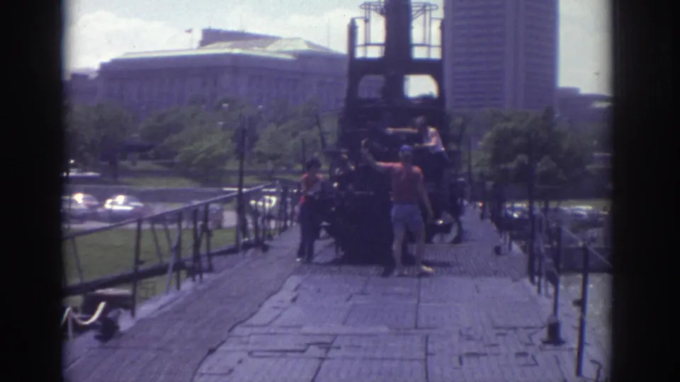 1974 Cleveland home-movie frame showing people on a wooden walkway near a dark structure with downtown buildings in the background.