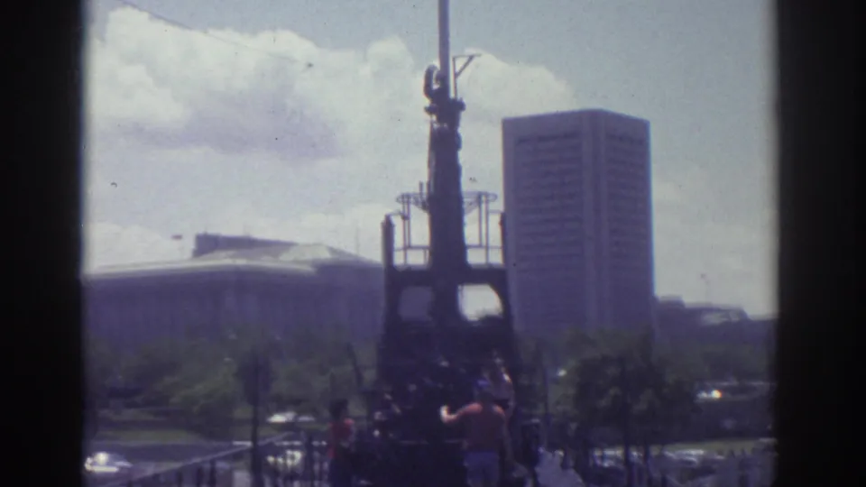 Cleveland 1974 home-movie view of a dark structure, people on a walkway, and high-rise buildings beyond a grassy area.