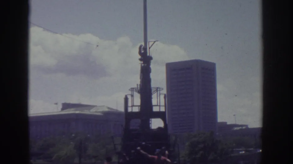 1974 Cleveland 8mm frame with a centered dark structure, people near its base, and a tall downtown building in the background.