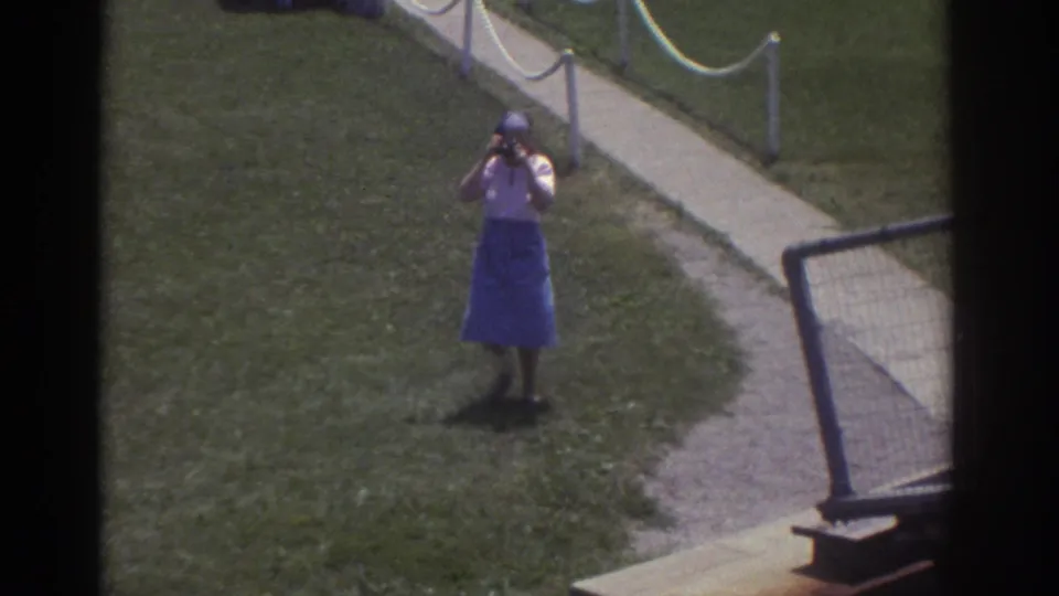 1974 Cleveland home-movie frame showing a woman photographing from grass near a paved path and rope-lined walkway.