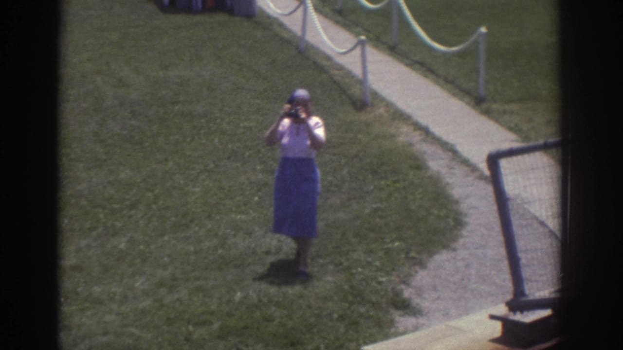 In Cleveland home-movie footage from 1974, a woman in a white blouse and blue skirt raises a camera while standing on grass near a path.
