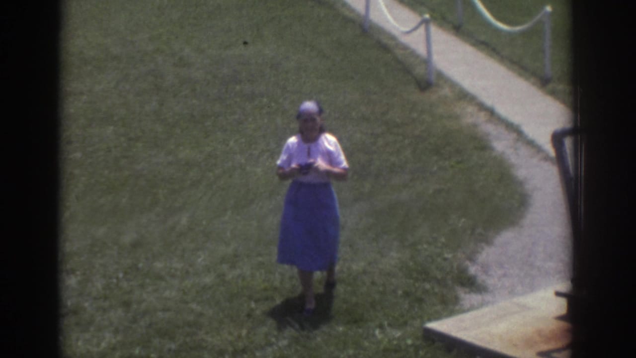Woman on a grassy lawn holding a camera below her face near a curved walkway.